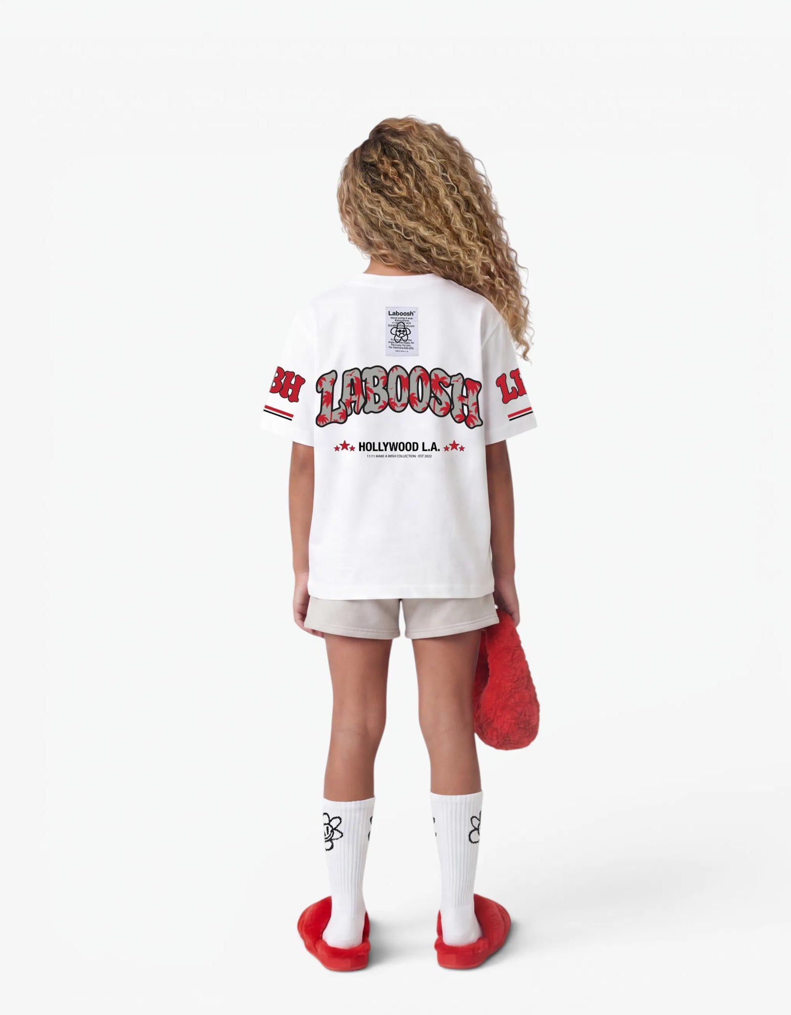 Girl wearing the white LBH oversized t-shirt, back view, showing the red Laboosh™ Hollywood L.A. logo print across the back, styled with matching grey shorts on a white background.