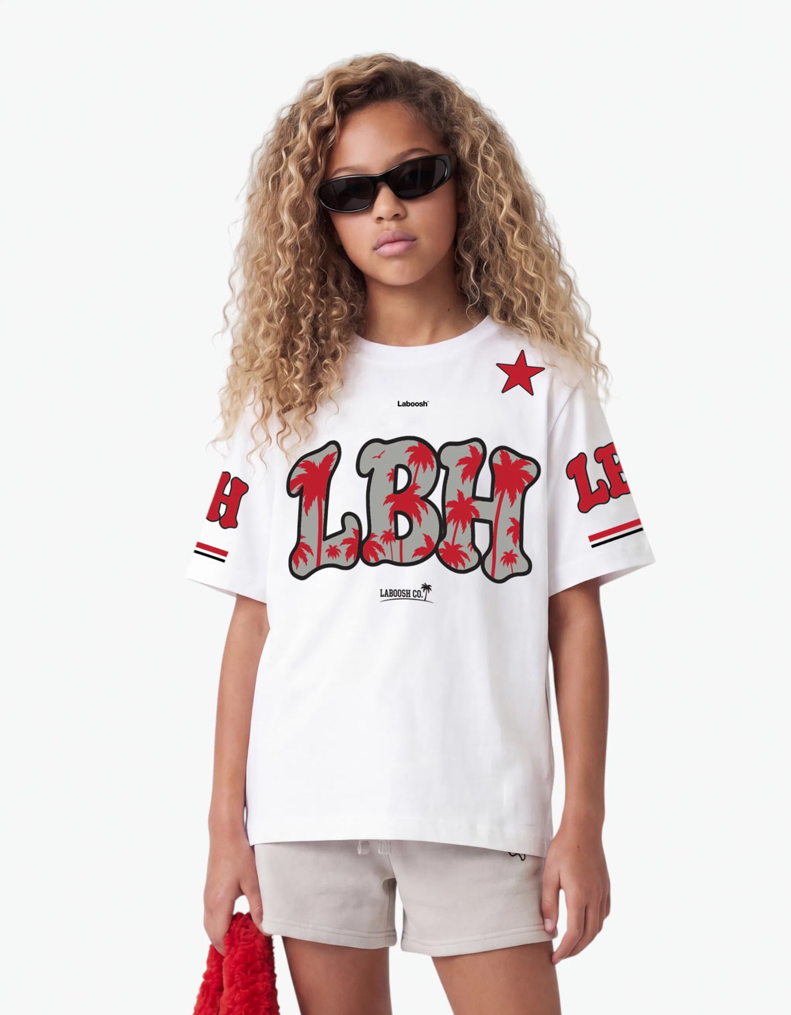 Girl in the white LBH oversized t-shirt, half-body close-up, highlighting the bold red LBH palm-tree graphic and Laboosh™ logo on the chest, styled with matching grey shorts on a white background.