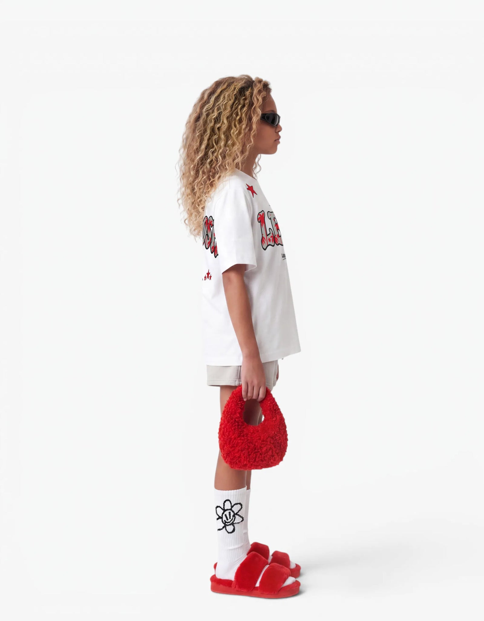 Girl wearing the white LBH oversized t-shirt with red palm-tree graphic, side view, showing the relaxed dropped-shoulder fit, styled with matching grey shorts on a white background.