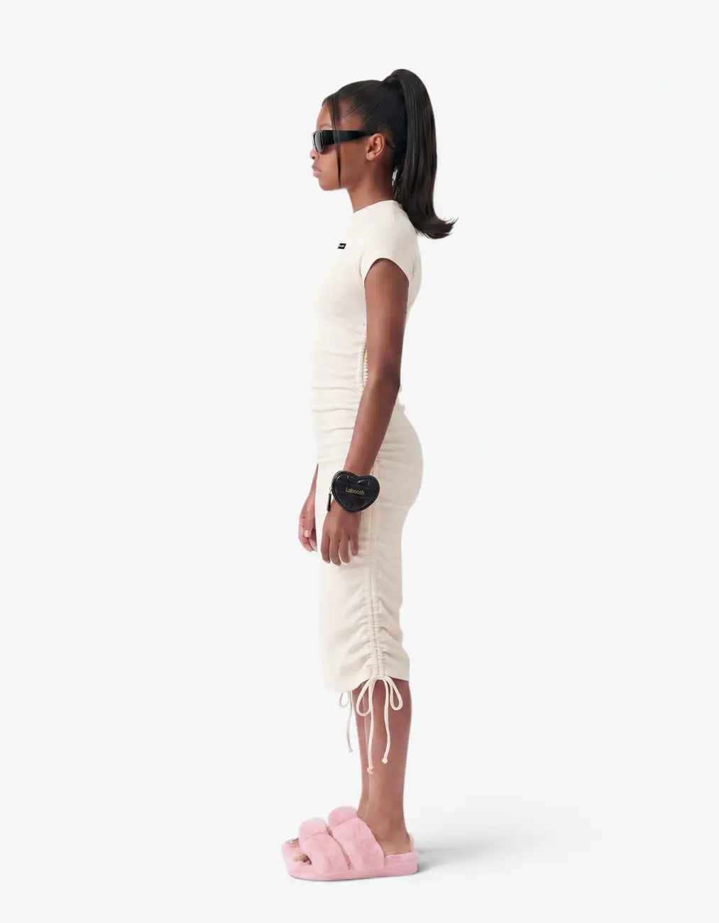 Side view of a girl wearing the cream ribbed ruched dress showing the ruched seam and side ties, on a white background