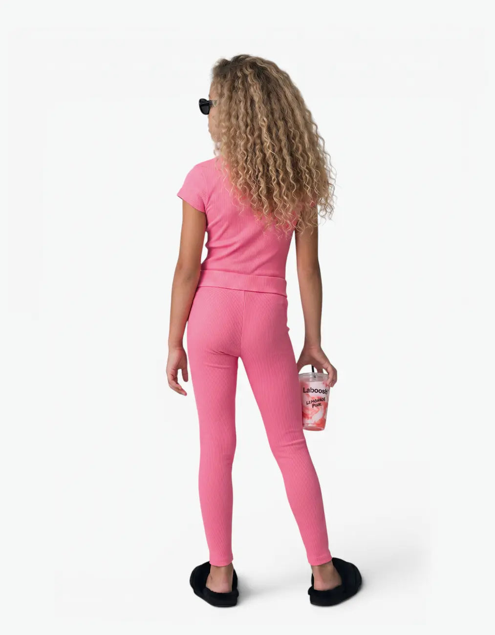Girl wearing pink ribbed short sleeve top, back view, with the Laboosh™ logo on the chest, on a white studio backdrop.